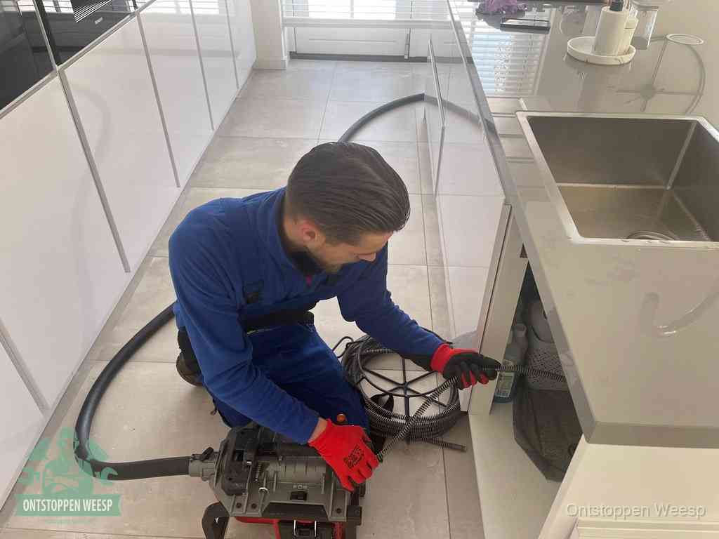 Een monteur ontstopt de keukenafvoer onder de spoelbak met een elektrische veermachine in Weesp