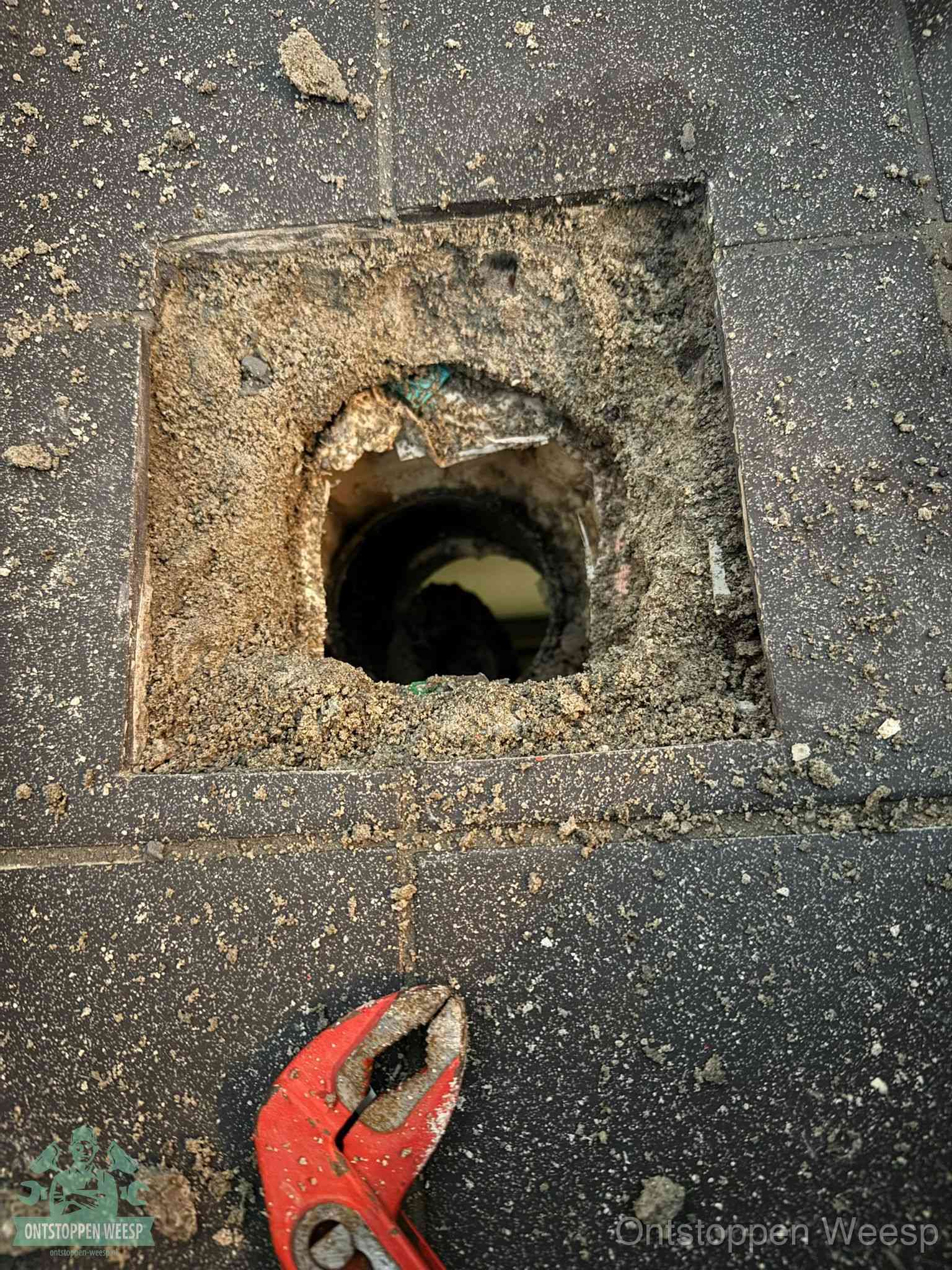 Een geopend rioolputje in een betontegelvloer met losgemaakt rooster en een zichtbare ronde afvoerbuis, het achterhalen waar de afvoerverstopping zit in Weesp