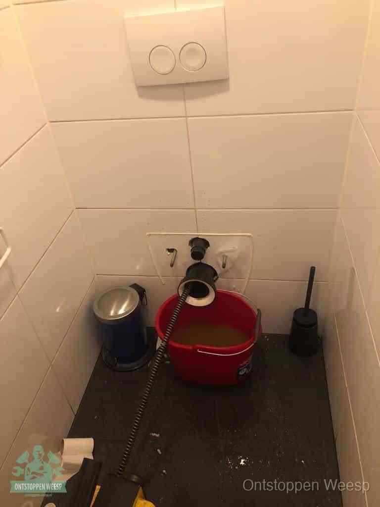 Een ontstopte wand-wc met verwijderde toiletpot waar een ontstoppingsveer in de wanduitgang verdwijnt, het achterhalen waar de verstopping zit in Weesp