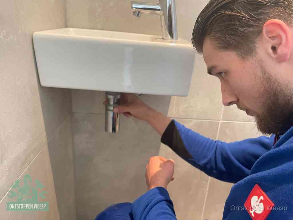 Een monteur draait met de hand de zwanenhalssifon onder een fonteinwasbak in de toiletruimte los om de bron van rioollucht in Weesp te vinden