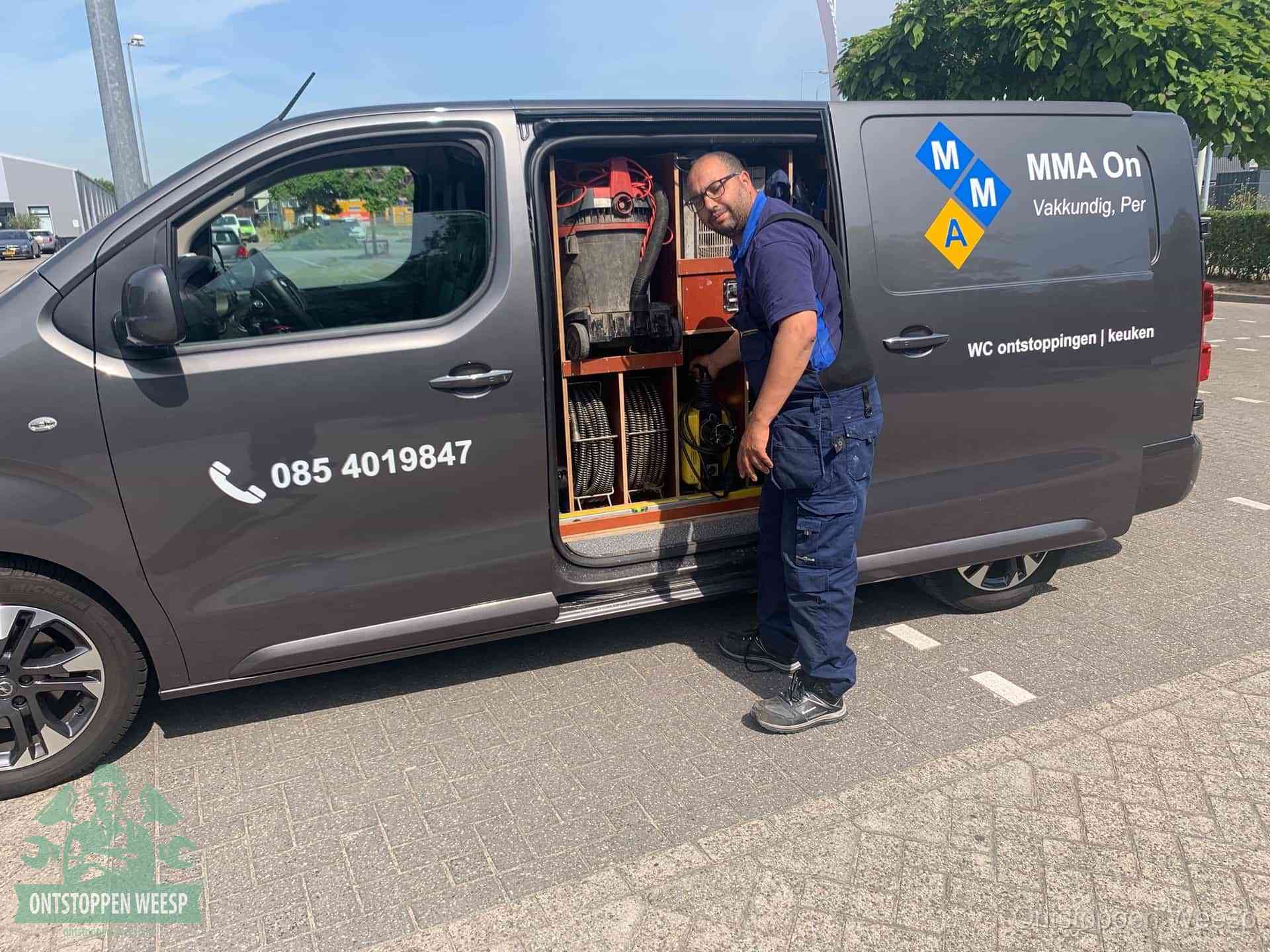 Een monteur bij de open schuifdeur van zijn bedrijfsbus op een parkeerplaats, klaar om de oorzaak van terugkerende afvoerverstoppingen aan te pakken in Weesp