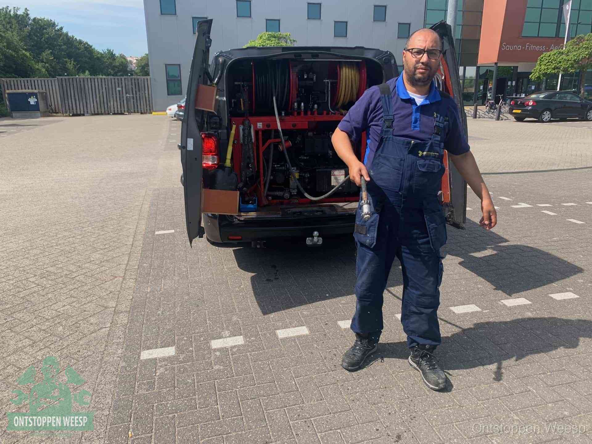 Een monteur bij zijn bestelbus met een hogedruk- en rioolreinigingsunit, klaar om de oorzaak van terugkerende wc-verstoppingen aan te pakken in Weesp