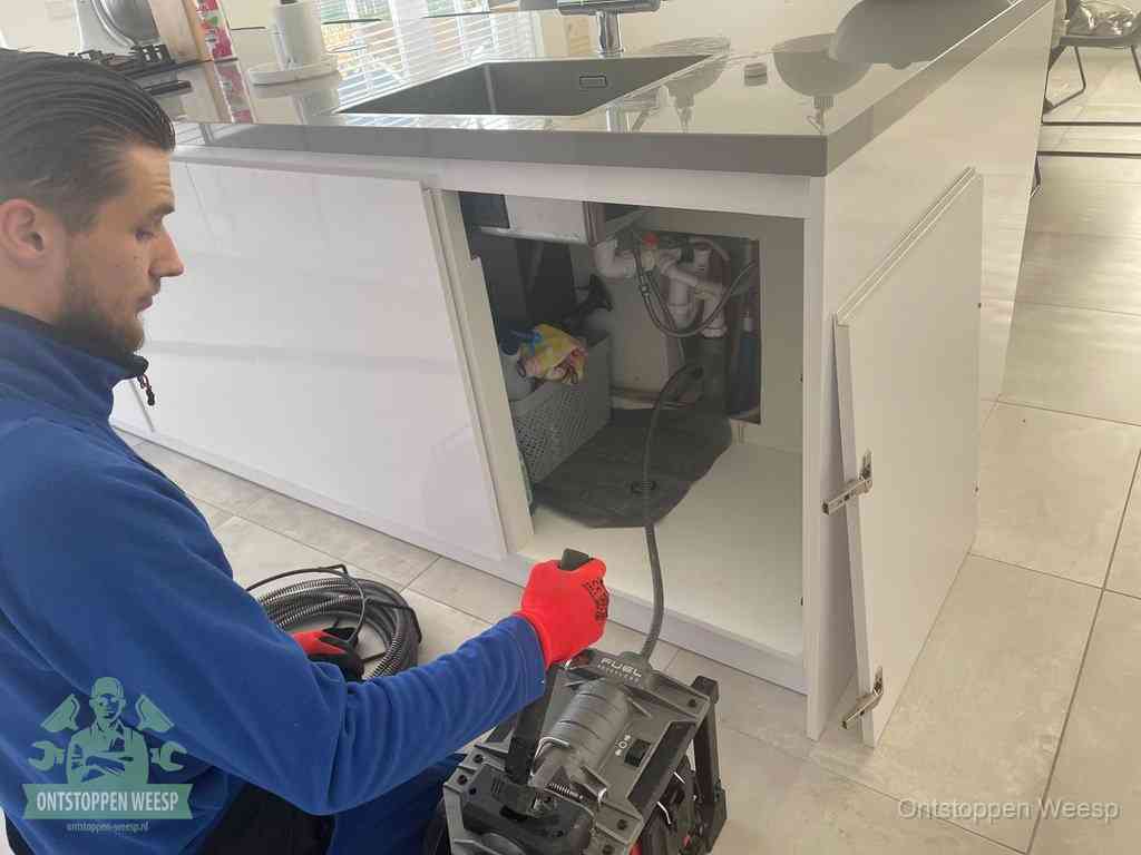 Monteur ontstopt met een veermachine een verstopte afvoer onder de gootsteen in Weesp