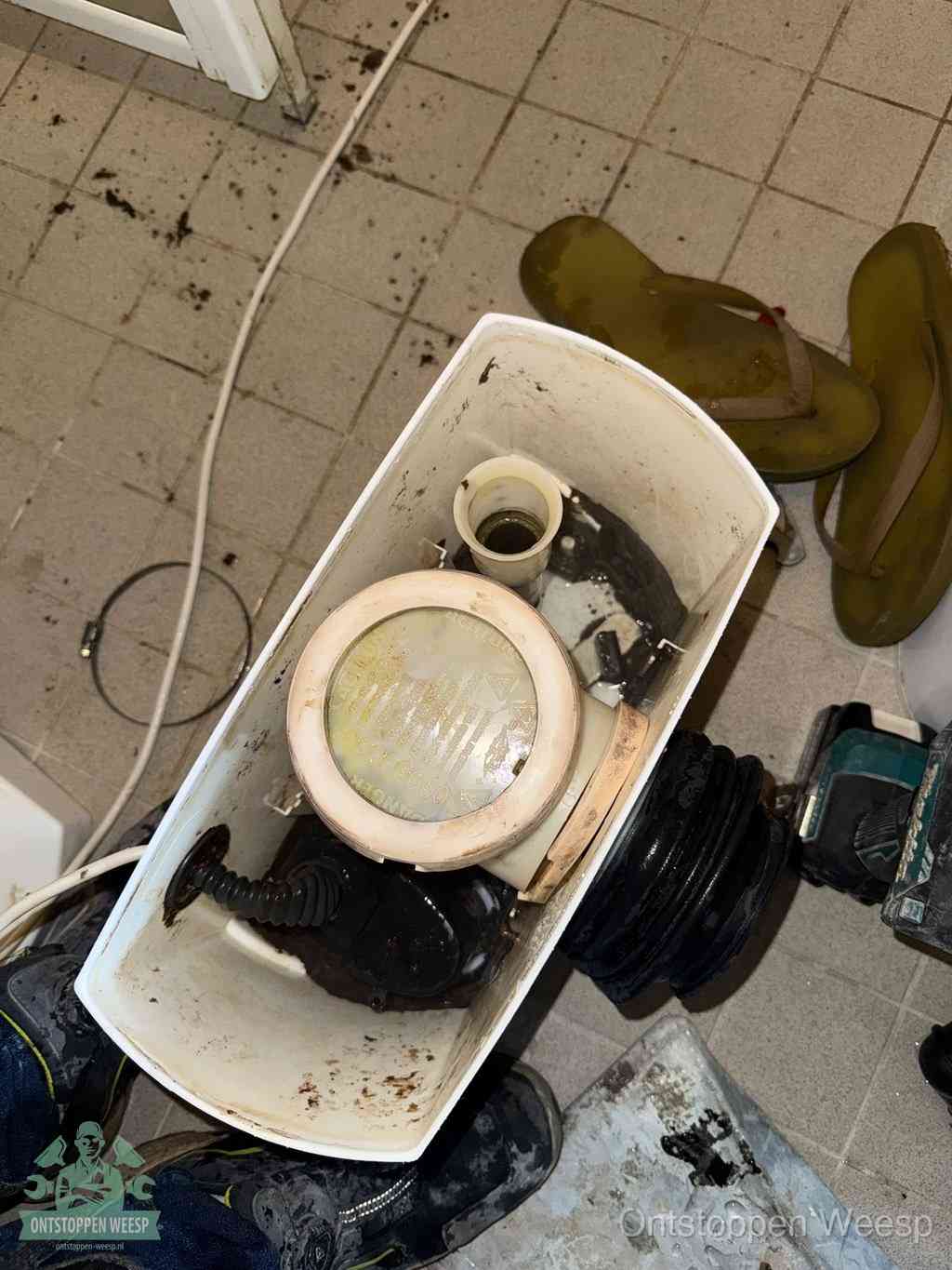 Een gedemonteerde toiletpot op de vloer met ontstoppingsveer en gereedschap ernaast, het verwijderen van een vastzittend voorwerp uit de wc in Weesp