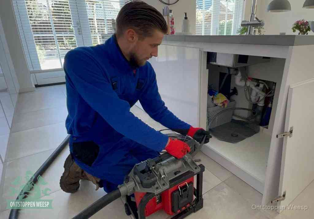 Een monteur voert een professionele elektrische ontstoppingsmachine in de afvoer onder de spoelbak tijdens het gootsteen ontstoppen in Weesp